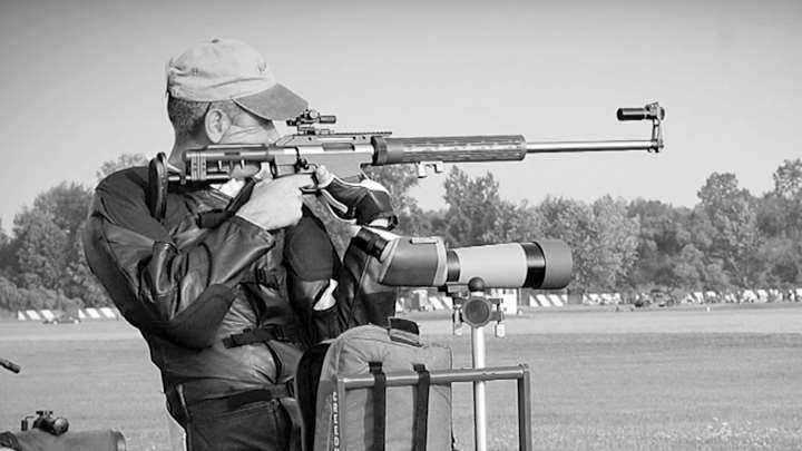 Black and white photo of Dennis Demille shooting with jacket, spotting scope and competitive gear.