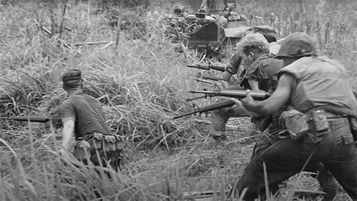 Marine stand ready with their M16s as a tank pushes forward through the brush.