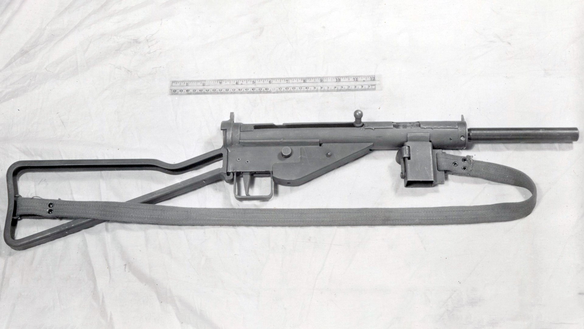 Right-side view of MP 3008 submachine gun with sling shown on white cloth with metal ruler for scale