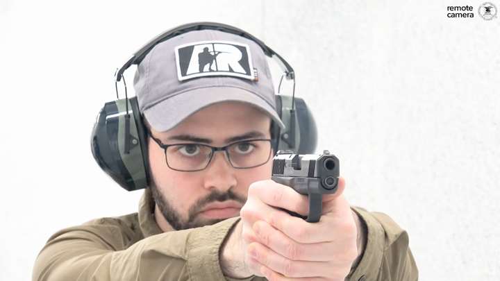 Man in protective shooting gear and wearing a ballcap shooting a black pistol on shooting range.