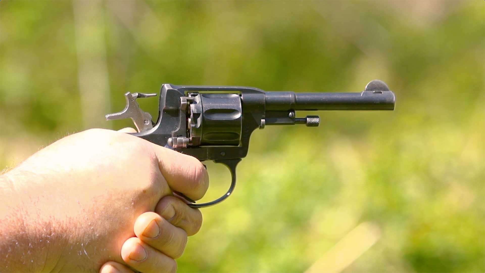 A pair of hands holding the M1895 Nagant revolver, aiming it on a green, outdoor range.