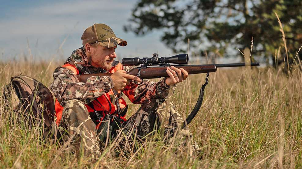 Man dressed in camouflage sitting in tall grass outdoors holding CZ 600 American bolt-action rifle with wood stock riflescope and sling