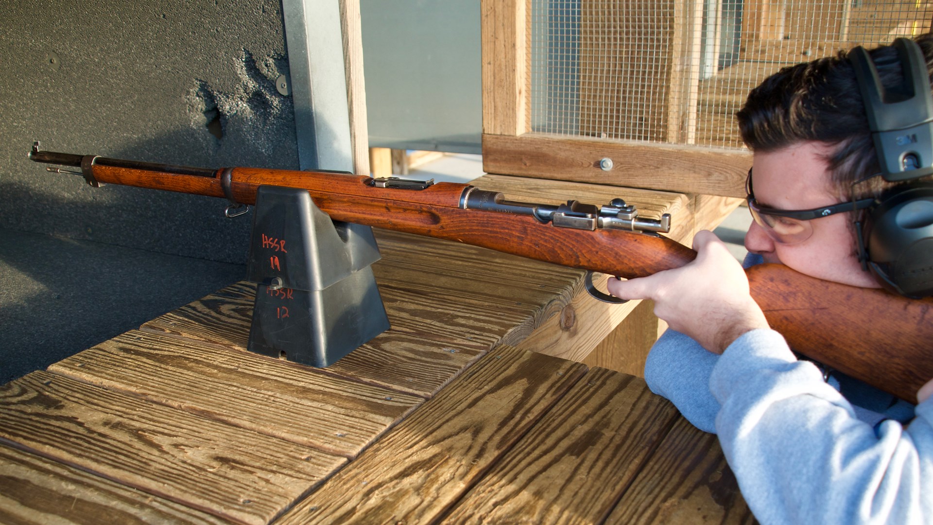 man sitting down at wood table holding swedish mauser bolt-action rifle shooting target range