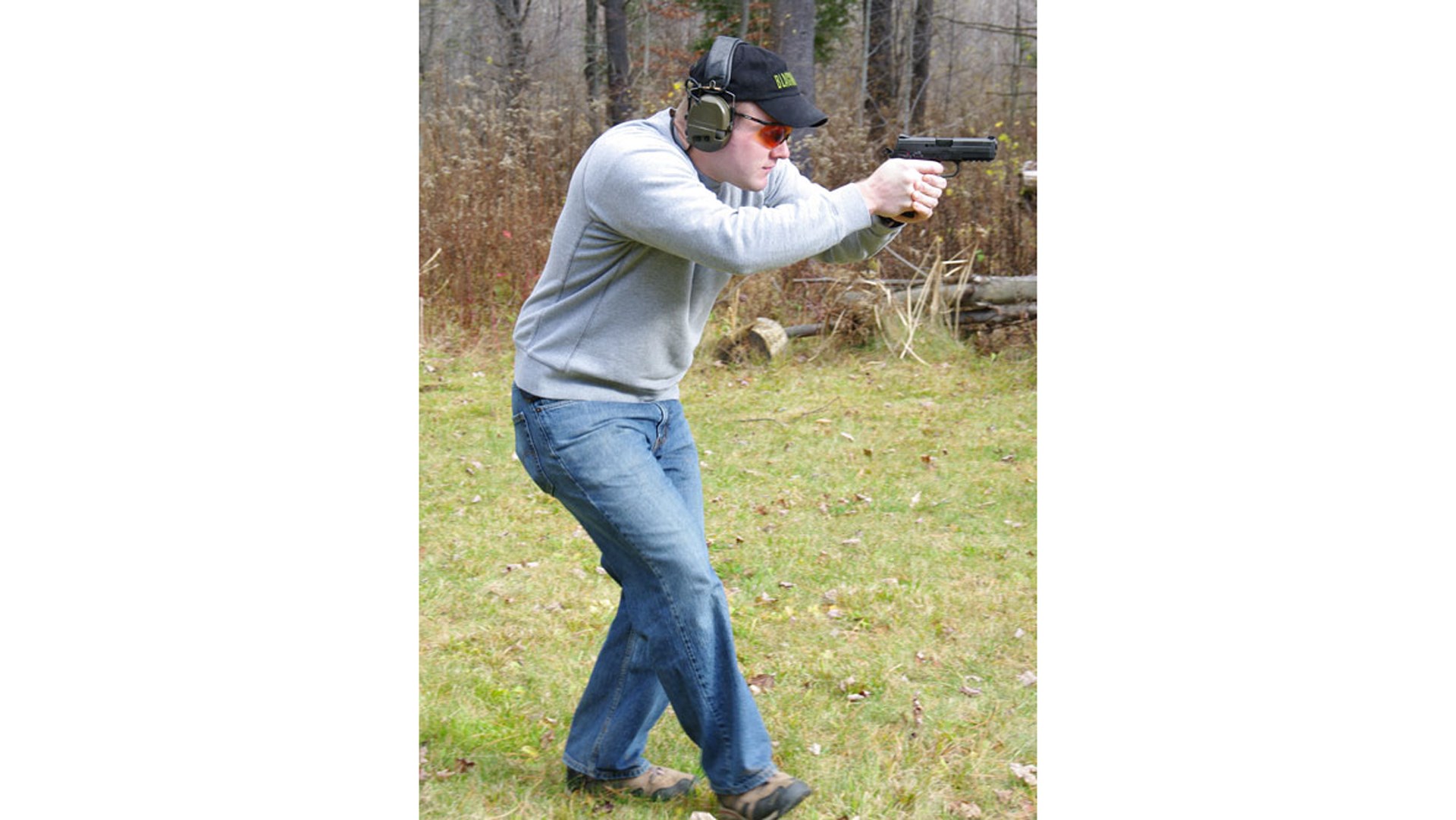 Man wearing jeans and gray longsleeve with ballcap and earmuffs walking while shooting handgun outdoors grass trees woods background