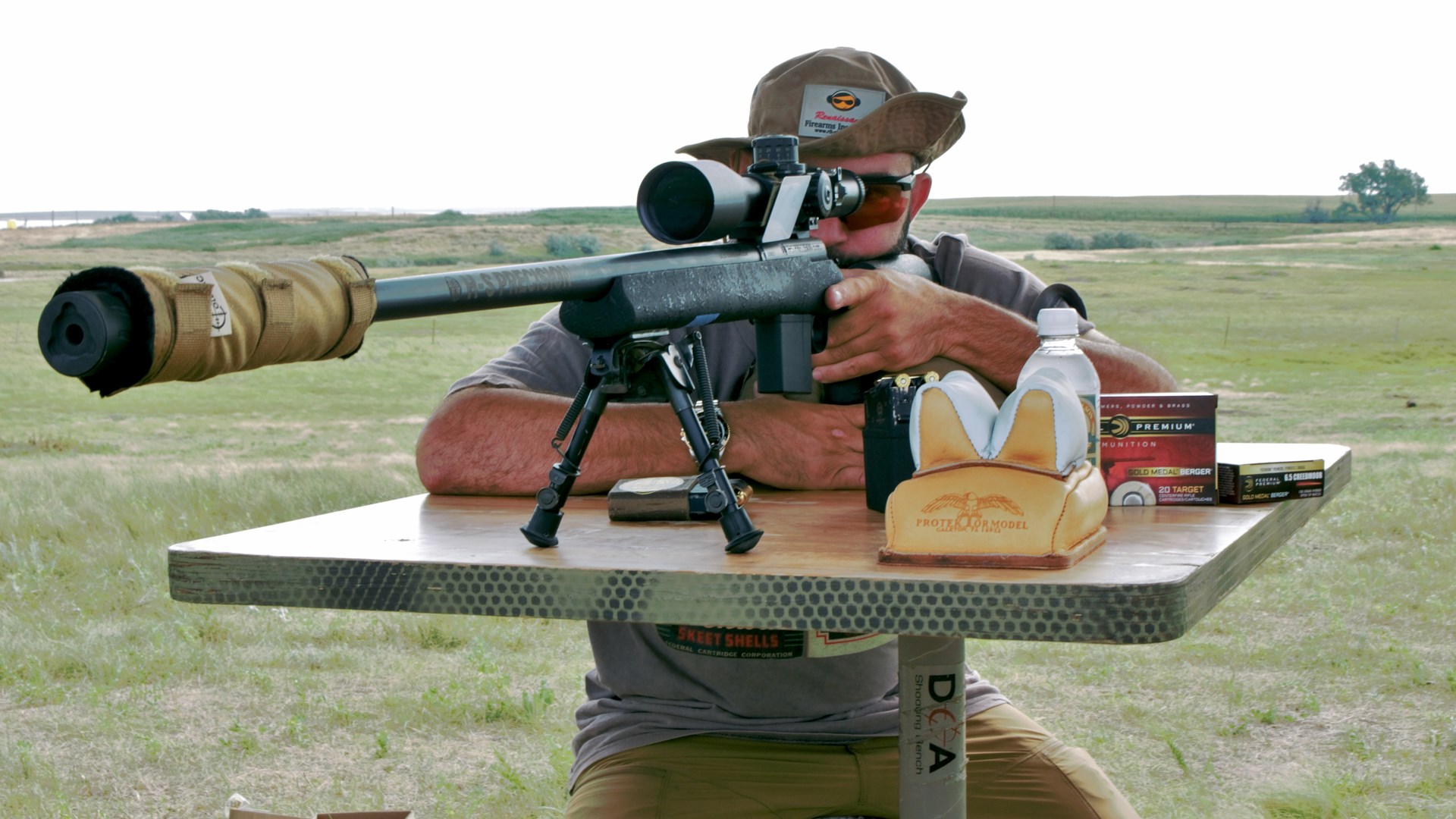 Author Frank Melloni wearing hat sunglasses sitting on shooting bench with suppressed bolt-action rifle left-hand hold ammunition outdoors bipod gun table