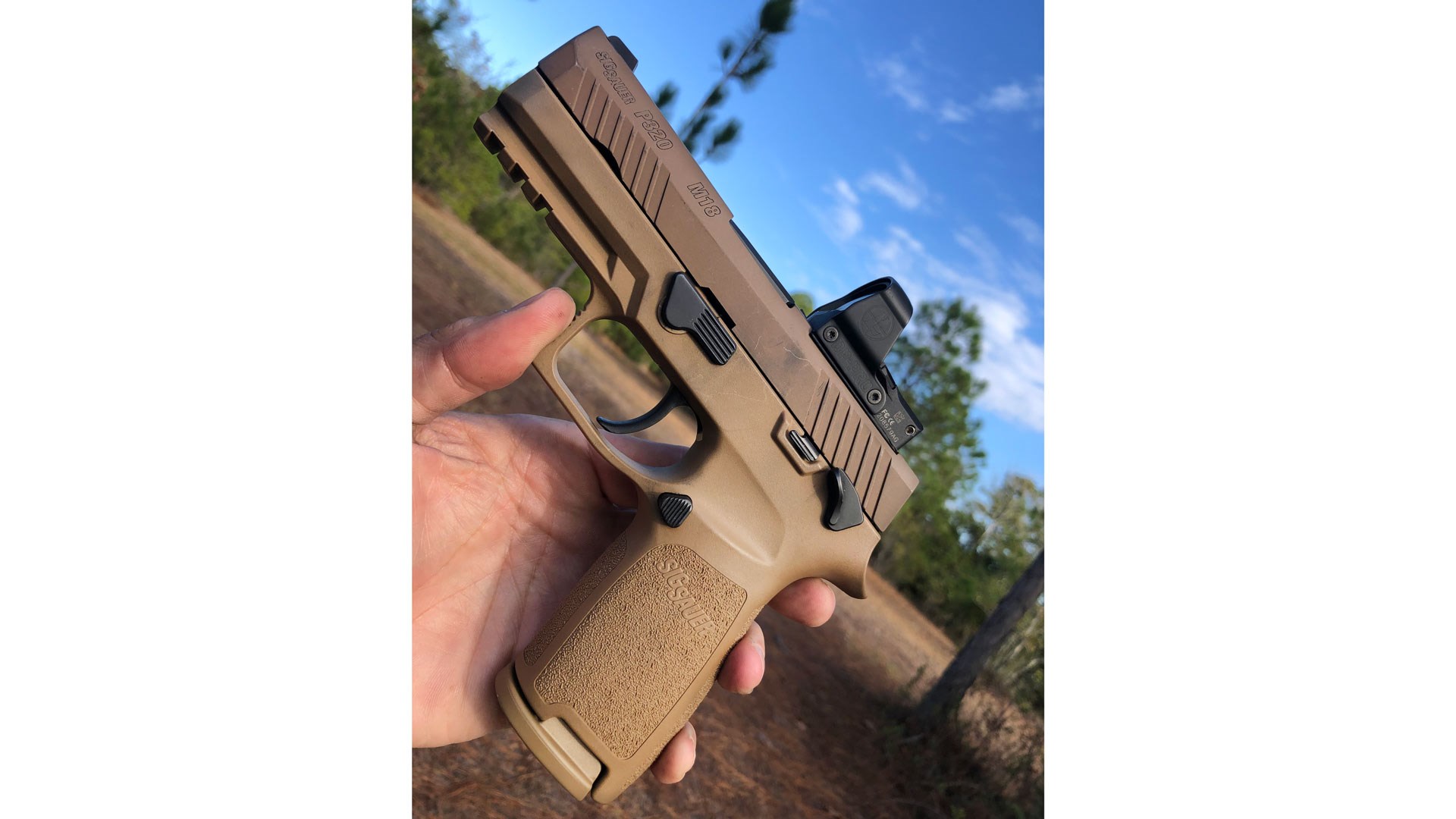 SIG Sauer M18 pistol in hand with optic outdoors blue sky trees