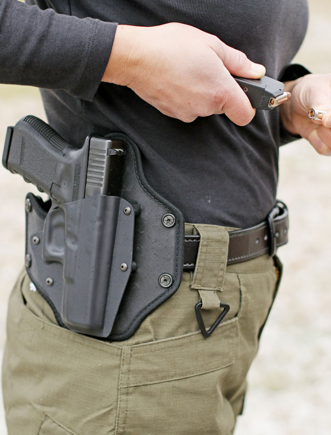Woman holding pistol magazine with Glock pistol in black outside-the-waistband holster on right hip.