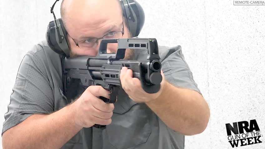Man wearing gray shirt earmuffs glasses shooting shotgun facing camera with text on image noting remote camera nra gun of the week