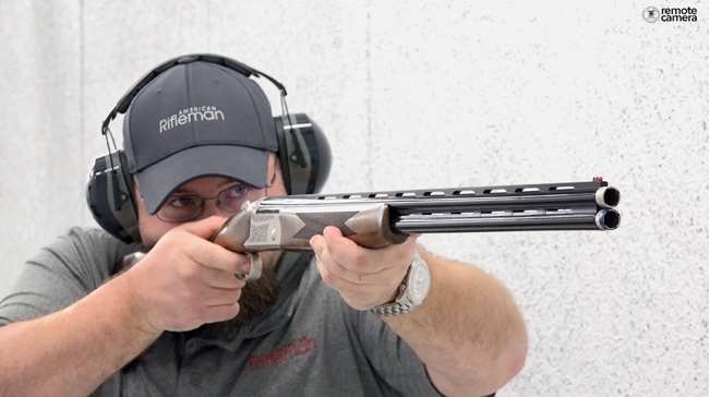 Man on shooting range wearing gray ballcap and t-shirt with American Rifleman embroidered and shooting a over-under shotgun.