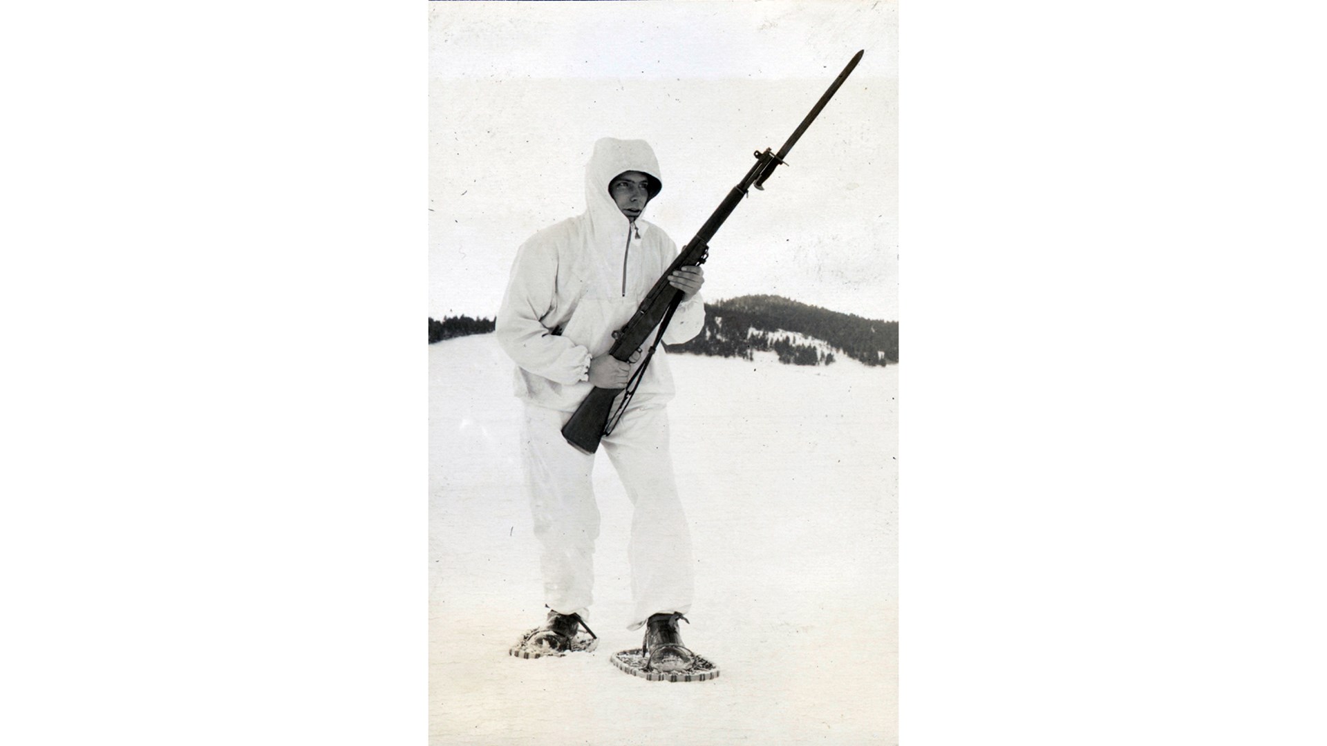 Man wearing white clothing snowshoes holding rifle in the snow mountain background
