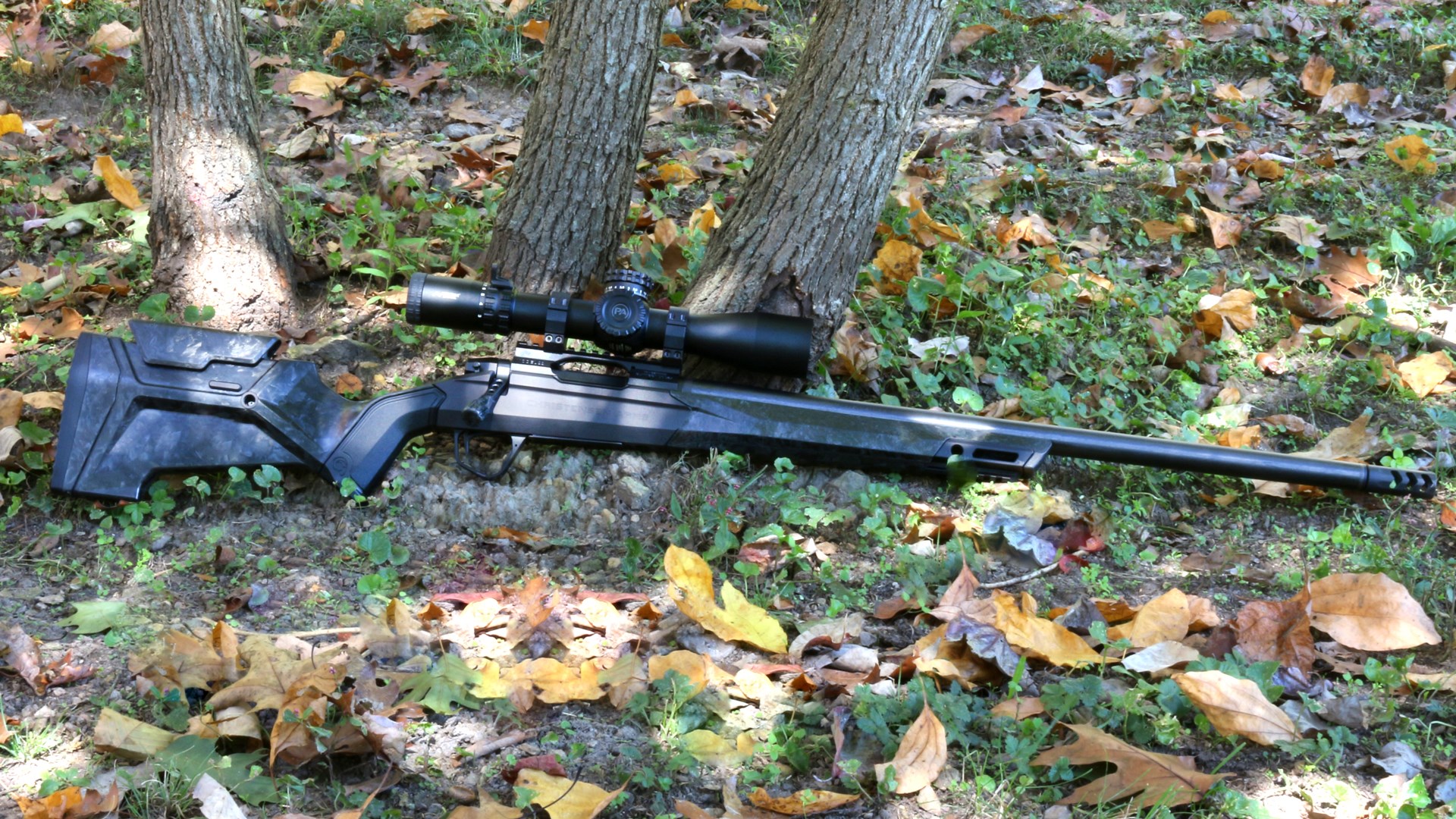 Right-side view of Christensen Arms Modern Hunting Rifle MHR bolt-action resting on ground base of three trees leaves outdoors