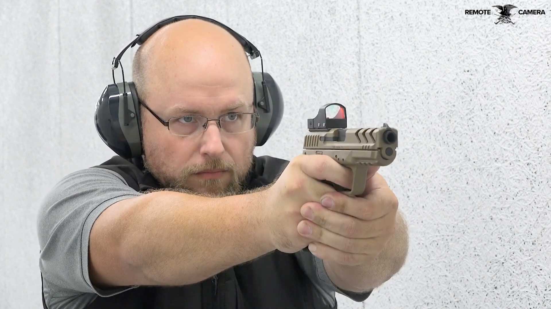 man wearing black vest gray shirt earmuffs glasses shooting brown pistol handgun