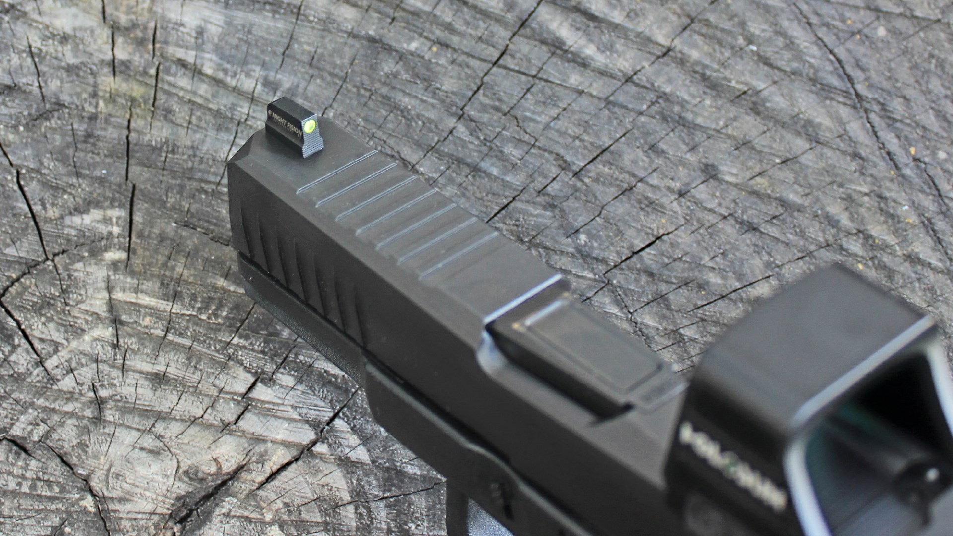 Top, forward view of Lone Wolf Timberwolf 19X slide with focus on Night Fision sight and slide serrations. Faded Holosun logo on red-dot optic. Wood stump background.