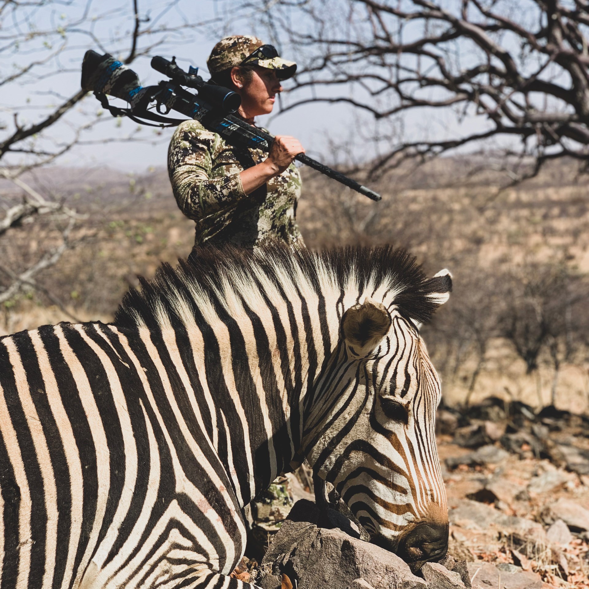 Female huntress in camouflage with rifle slung on shoulder looking into distance behind zebra laying on ground after hunting