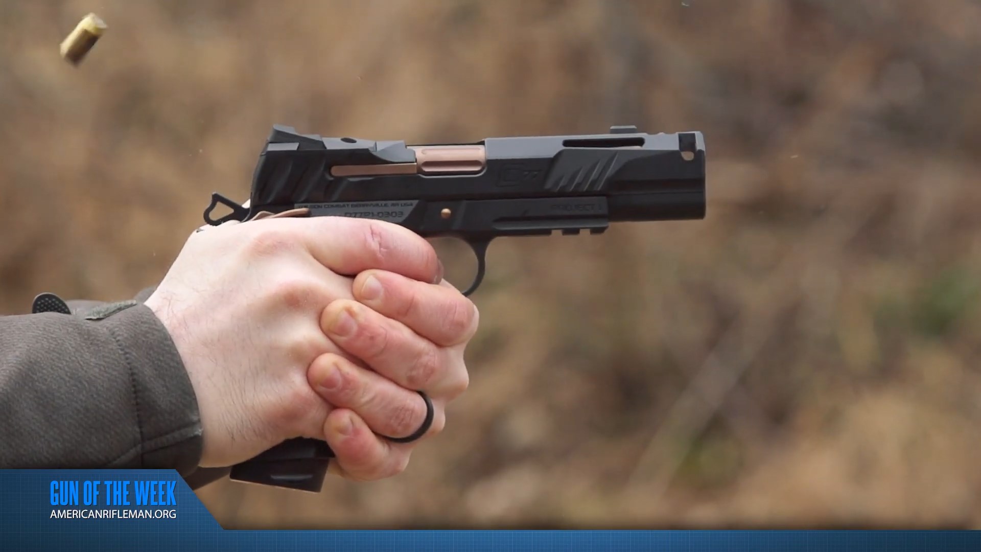 AMERICAN RIFLEMAN GUN OF THE WEEK video screenshot of man's hand shooting black wilson combat division 77 project 1 pistol with 9 mm shell casing flying through air green brush background outdoors