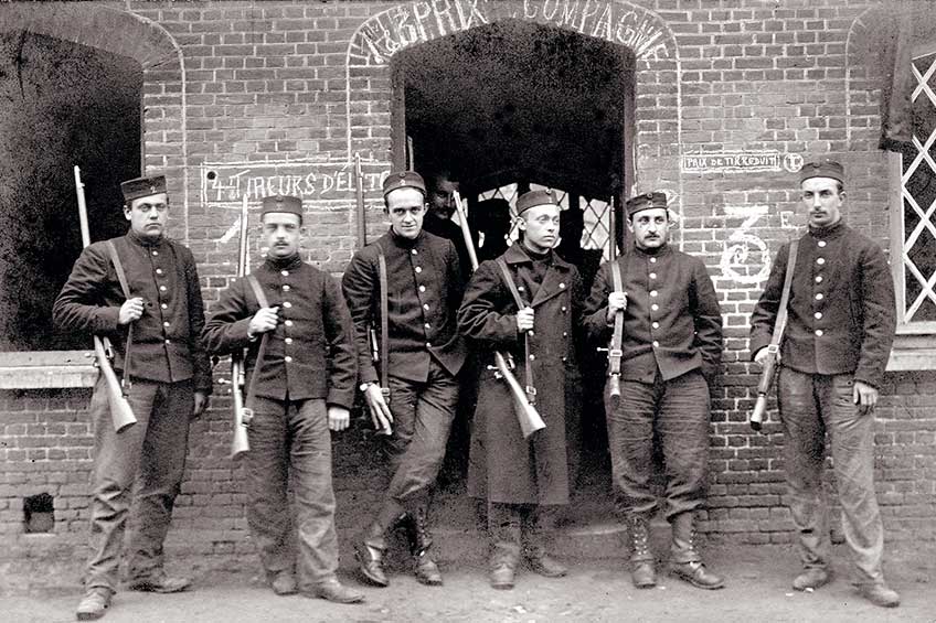 Black and white image of six soldiers in uniform with rifles slung on shoulders and brick background.