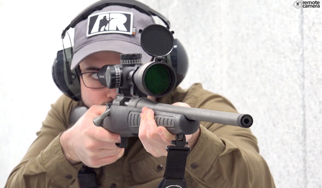Foward-facing view of rifleman with gray ballcap and green shirt shooting rifle in white indoor shooting range.