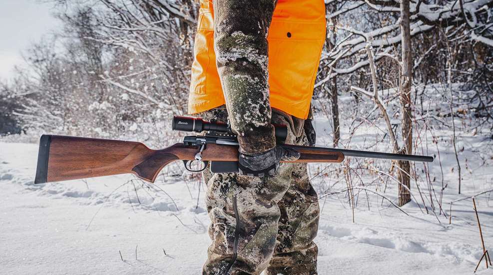 An orange-jacketed hunter walks through snowy woods carrying a wood-stocked Savage 334 bolt-action rifle at his side.