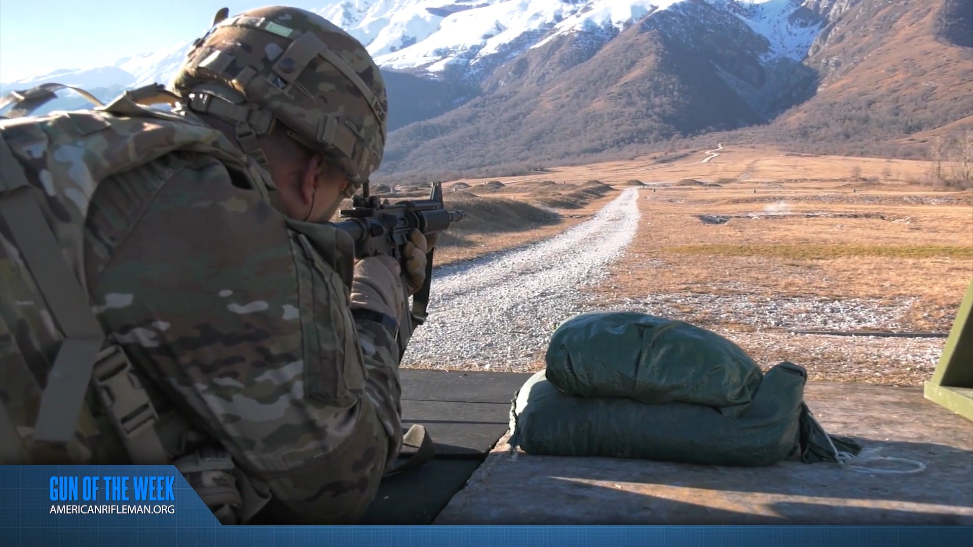 GUN OF THE WEEK AMERICANRIFLEMAN.ORG text banner beneath soldier with M4 firiing prone with gravel road heading to mountains with snow atop
