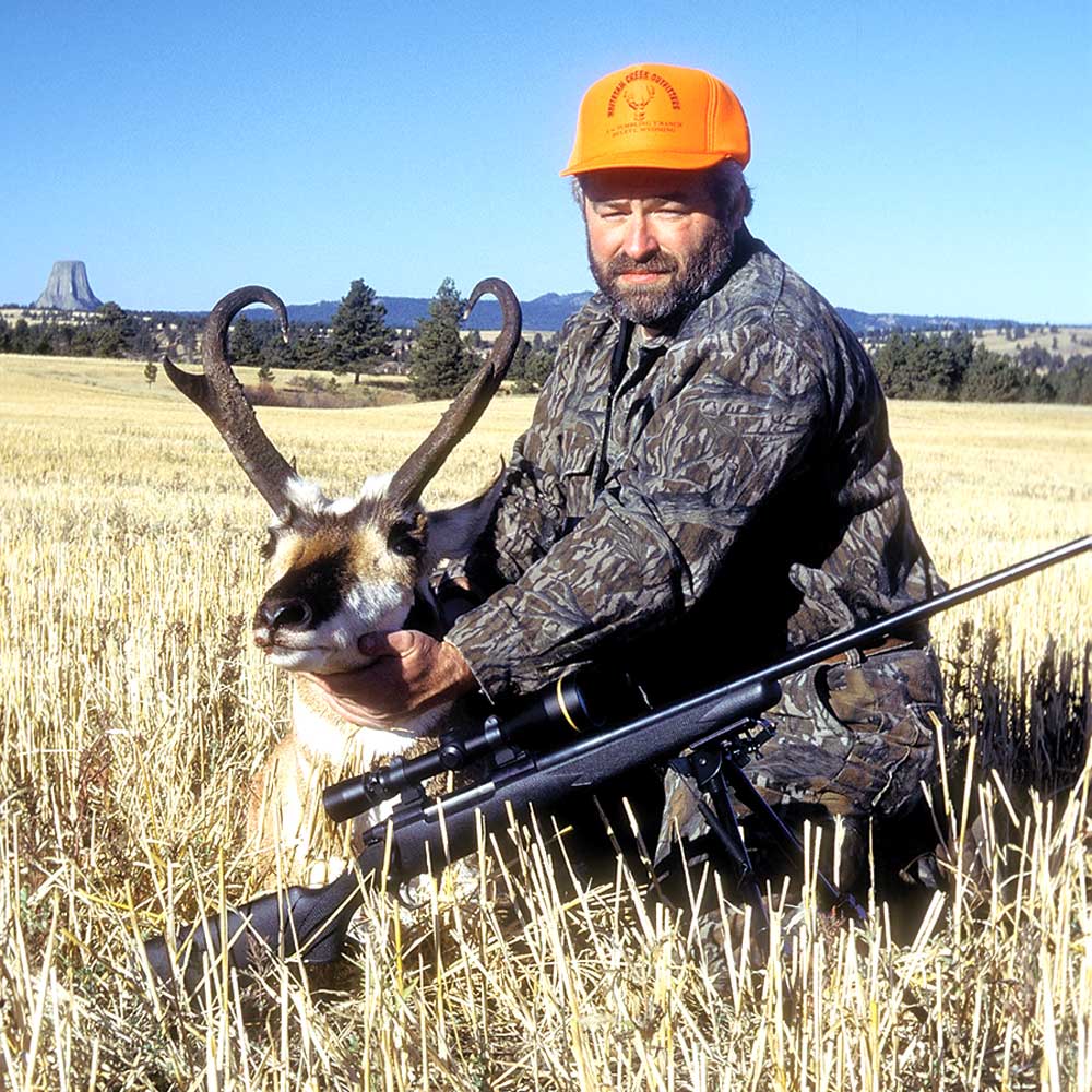 outdoors grass field blue sky devils tower mountain background hunter foreground antelope rifle orange hand camouflage clothing