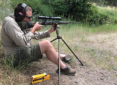 Thomas Haviland sitting and shooting bolt-action rifle on tripod support with yellow box of SIG Sauer ammunition.