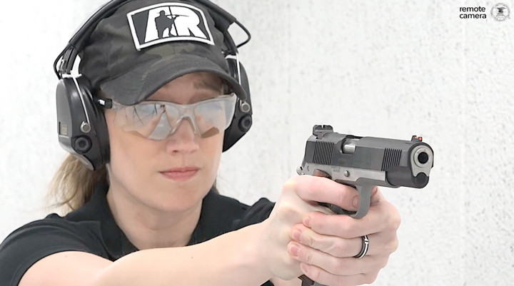 Lady with camo ballcap on and protective shooting gear on a white shooting range facing a remote camera shooting a two-tone springfield pistol.
