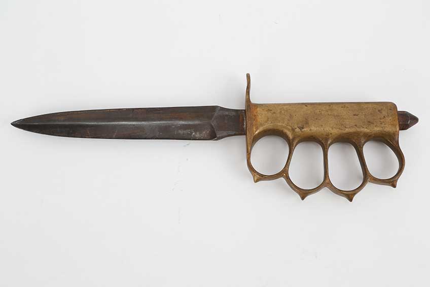 Left-side view of U.S. 1918 fighting knife on white background. Knife handle made of brass with guards for each finger.