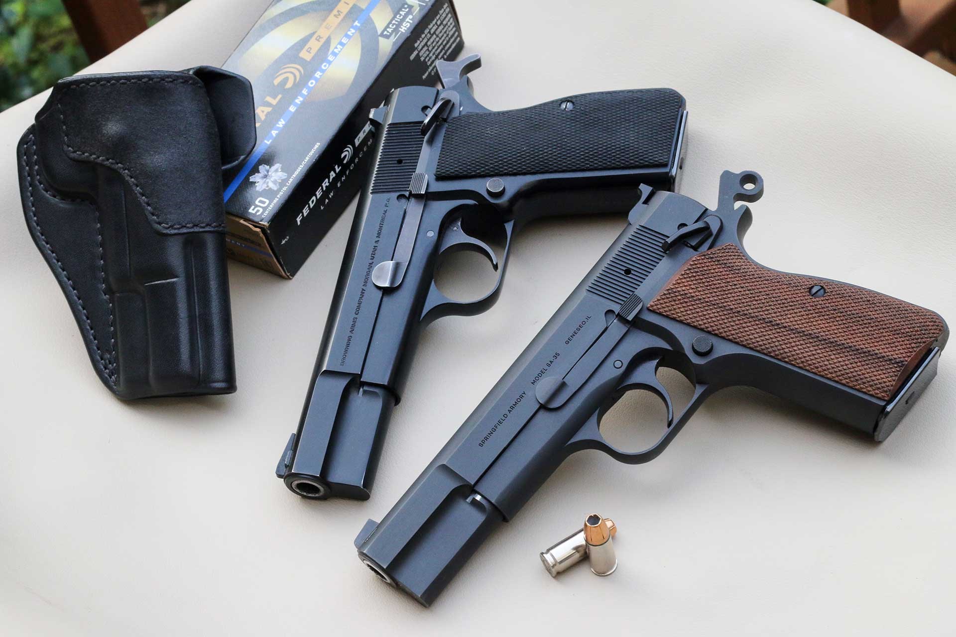 The new Springfield SA-35 High Power shown on a white table next to an original Browning Mk III Hi Power alongside a leather holster and several 9mm cartridges.