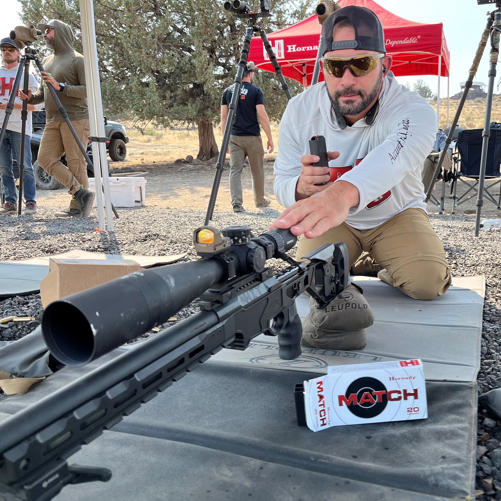 Man kneeling behind bolt-action seekins precision rifle with hornady ammunition box tent men standing background