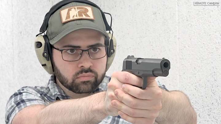 Man wearing protective shooting gear and a ballcap is firing a 1911 pistol on a shooting range.