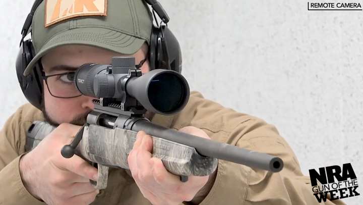 Man wearing a ballcap and protective shooting gear is shown shooting a camouflaged rifle.