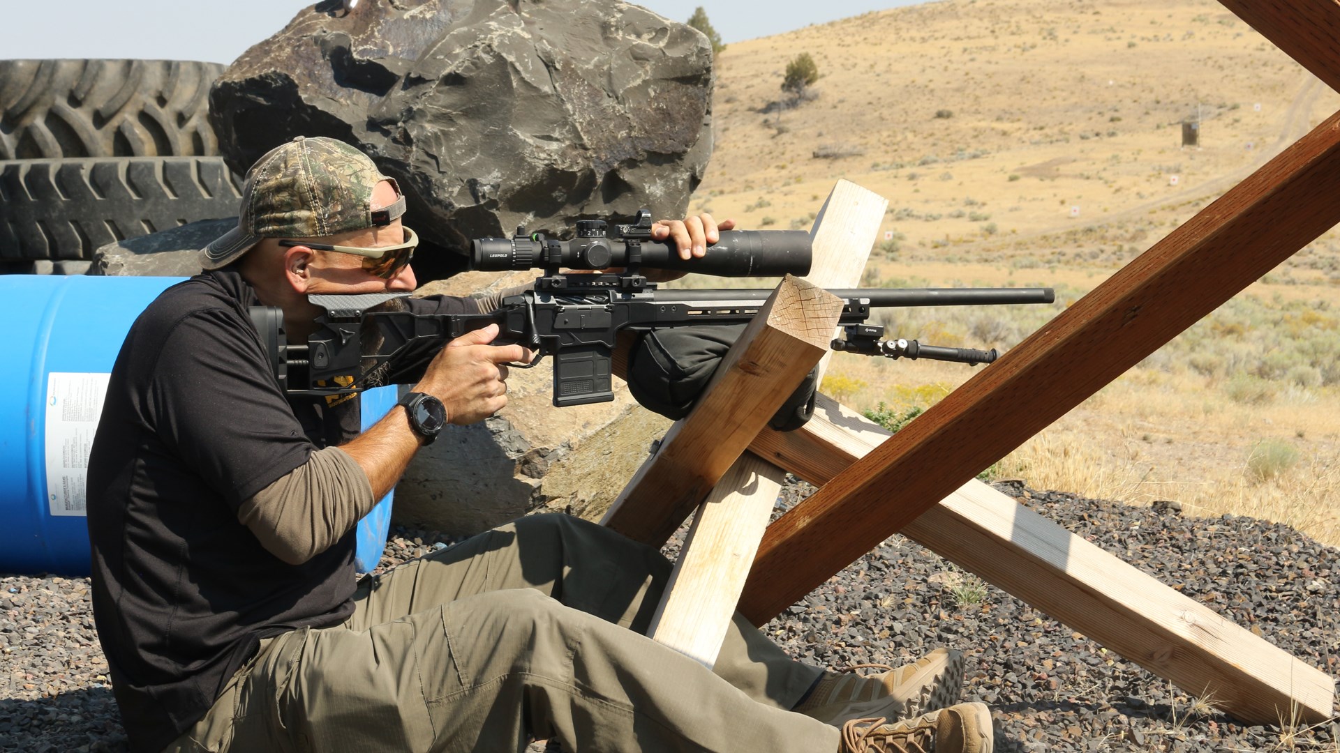 man behind rifle sitting on gravel near rocks tires blue drum outdoors resting rifle on barricade tank trap wood 4x4