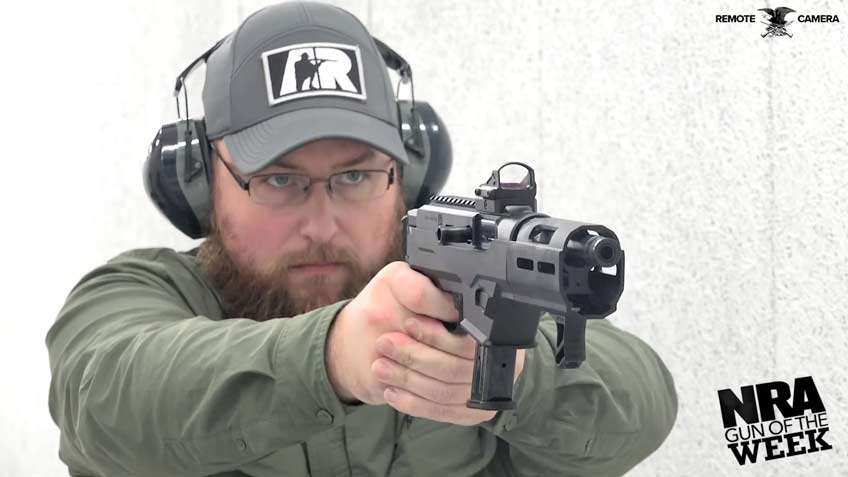 Man wearing a green shirt ballcap ear protection and glasses holding and shooting black pistol with text on image noting &quot;remote camera nra gun of the week&quot;