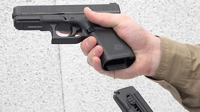 Man with green shirt holding Glock G44 rimfire pistol in right hand and inserting black magazine with opposing hand shown on white shooting range.