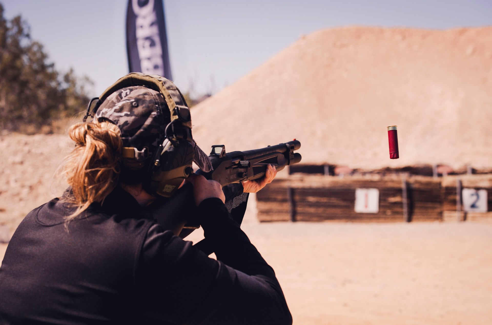 Woman backside view of upper torso long hair camo hat earmuffs outdoors shooting black mossberg shotgun with red shell captured in flight leaving the gun ejection dirt backgroud