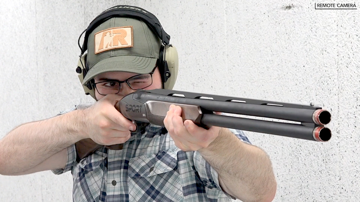Man wearing ballcap and protective shooting gear shooting a shotgun pointed toward camera.