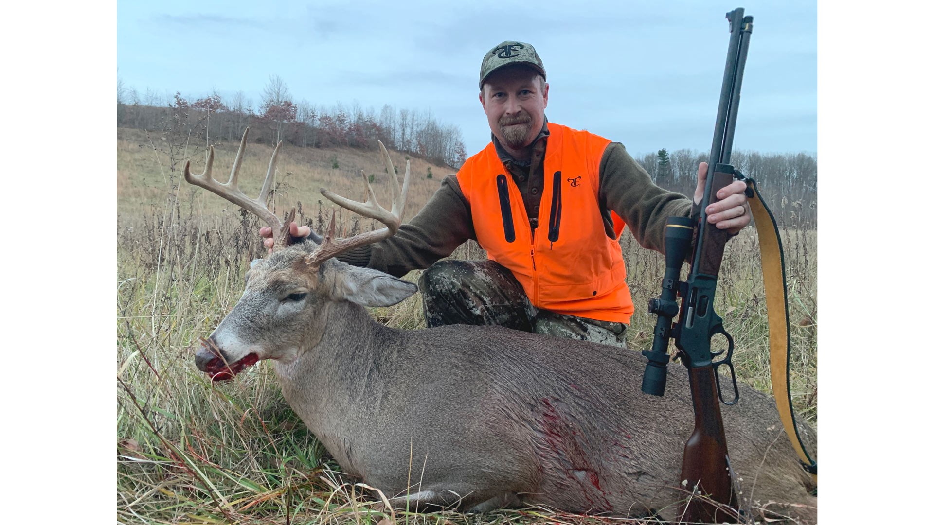 Man wearing orange vest with Henry Lever-Action rifle and whitetail deer outdoors 360 Buckhammer Remington hunter field grass treeline