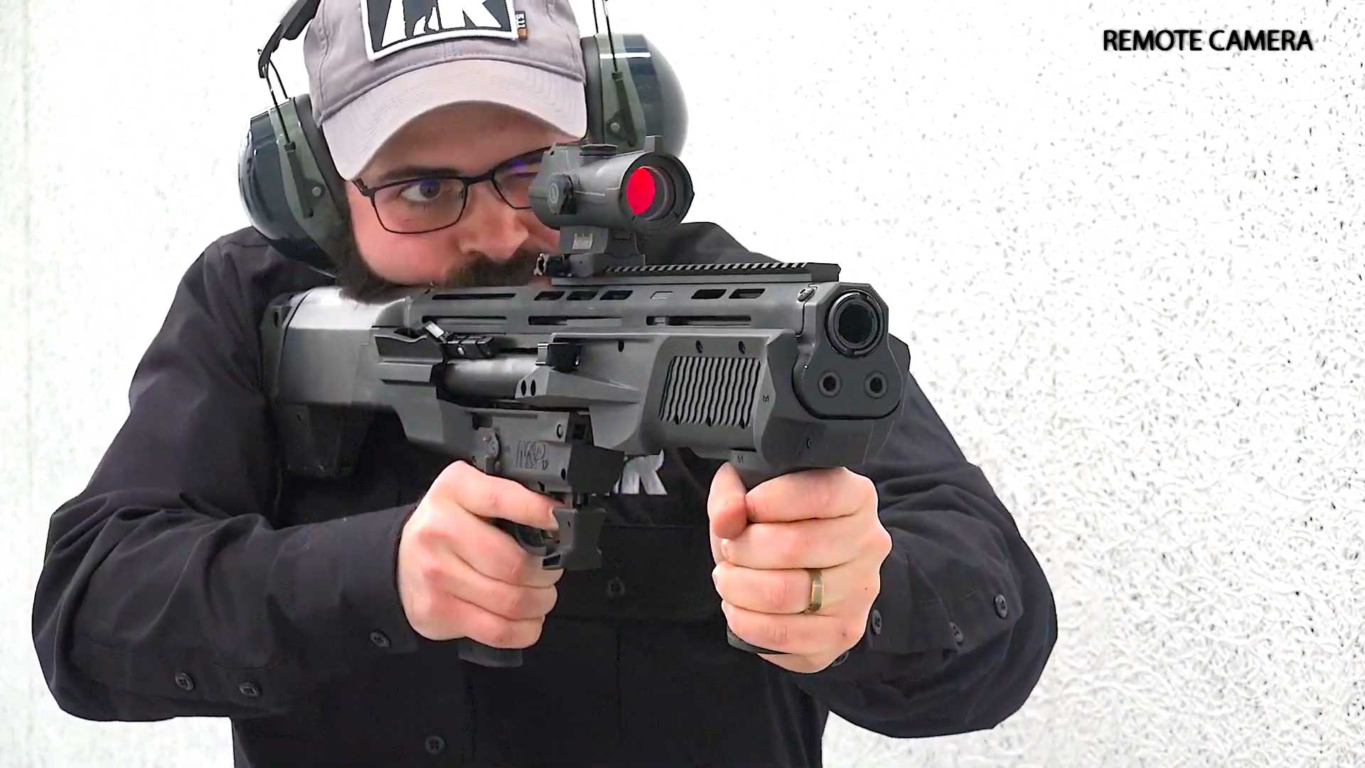 Man wearing black shirt hat earmuffs shooting black shotgun range indoors white walls remote camera