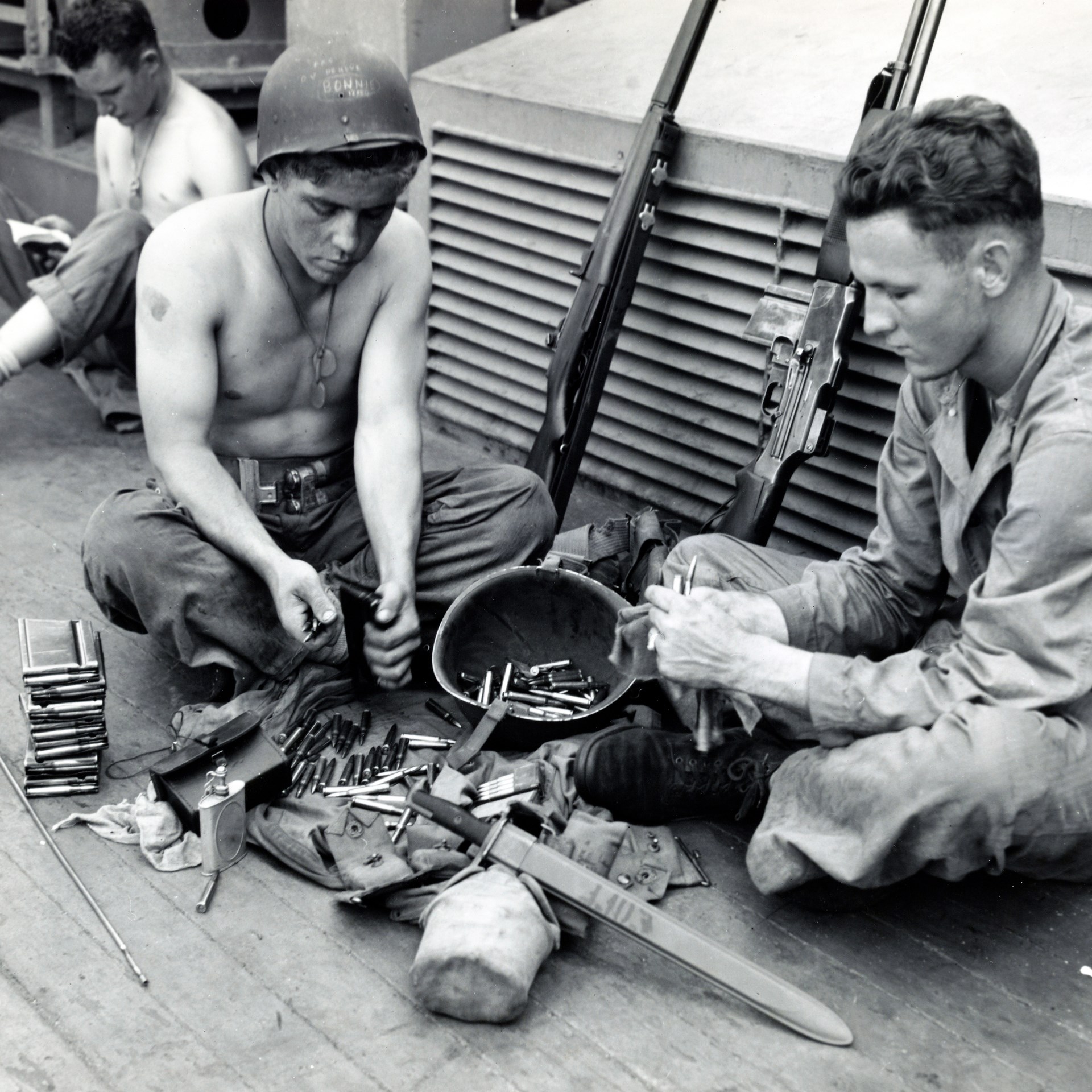Soldiers sitting while loading BAR magazines with ammunition rilfes leaned agains grate behind
