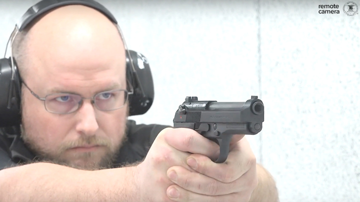Man wearing ear and eye protection shooting a Beretta pistol on a white shooting range.