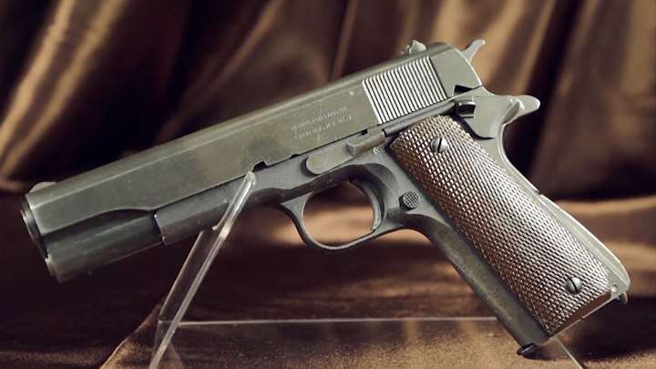 Left-side view of a Remington Rand M1911A1 on clear stand with drapes behind.