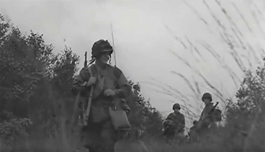 Black and white war-time photo of U.S. Paratroopers walking with gear along hedgerow and field.