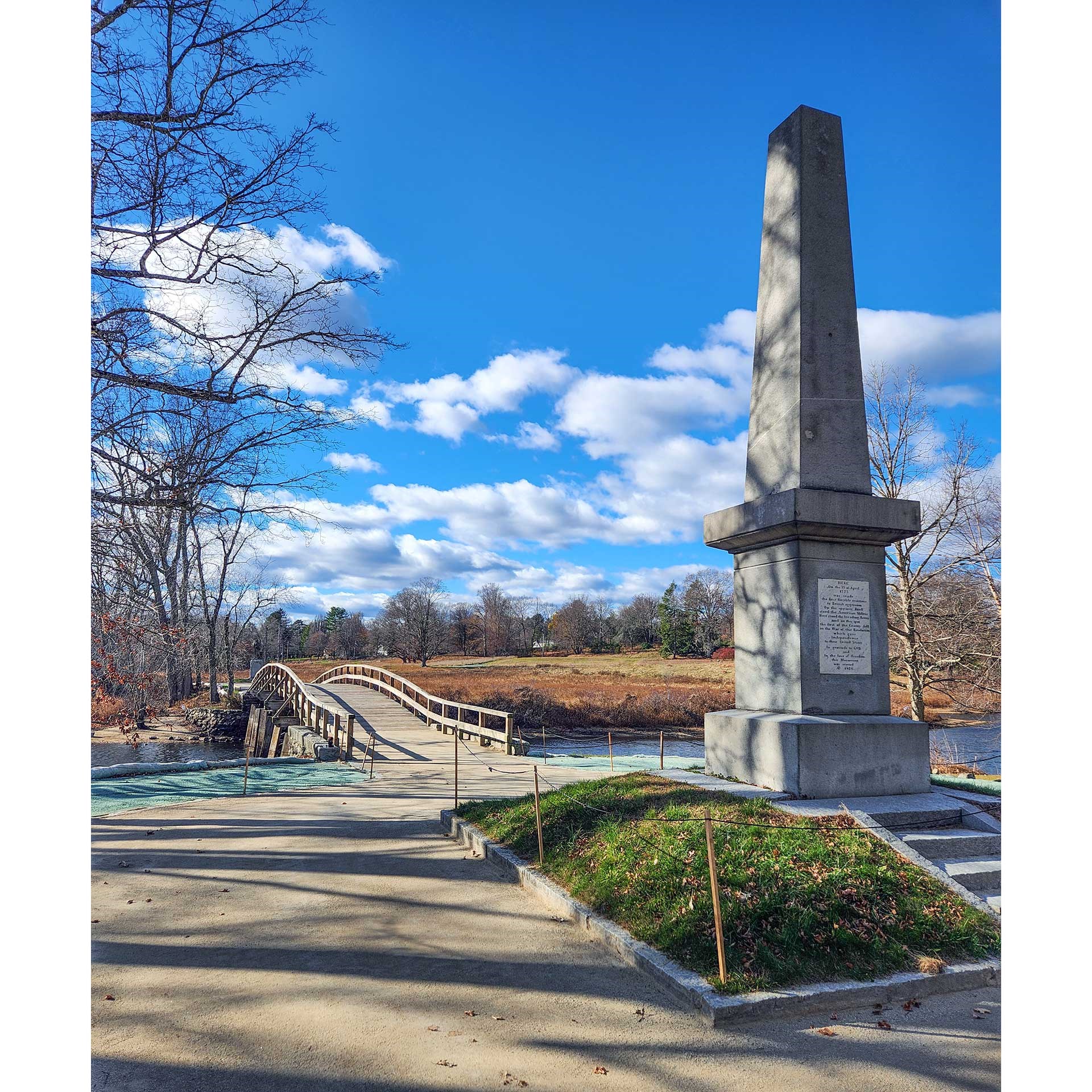 A wooden bridge shown next to a stone obelisk against a clear blue sky with white clouds.