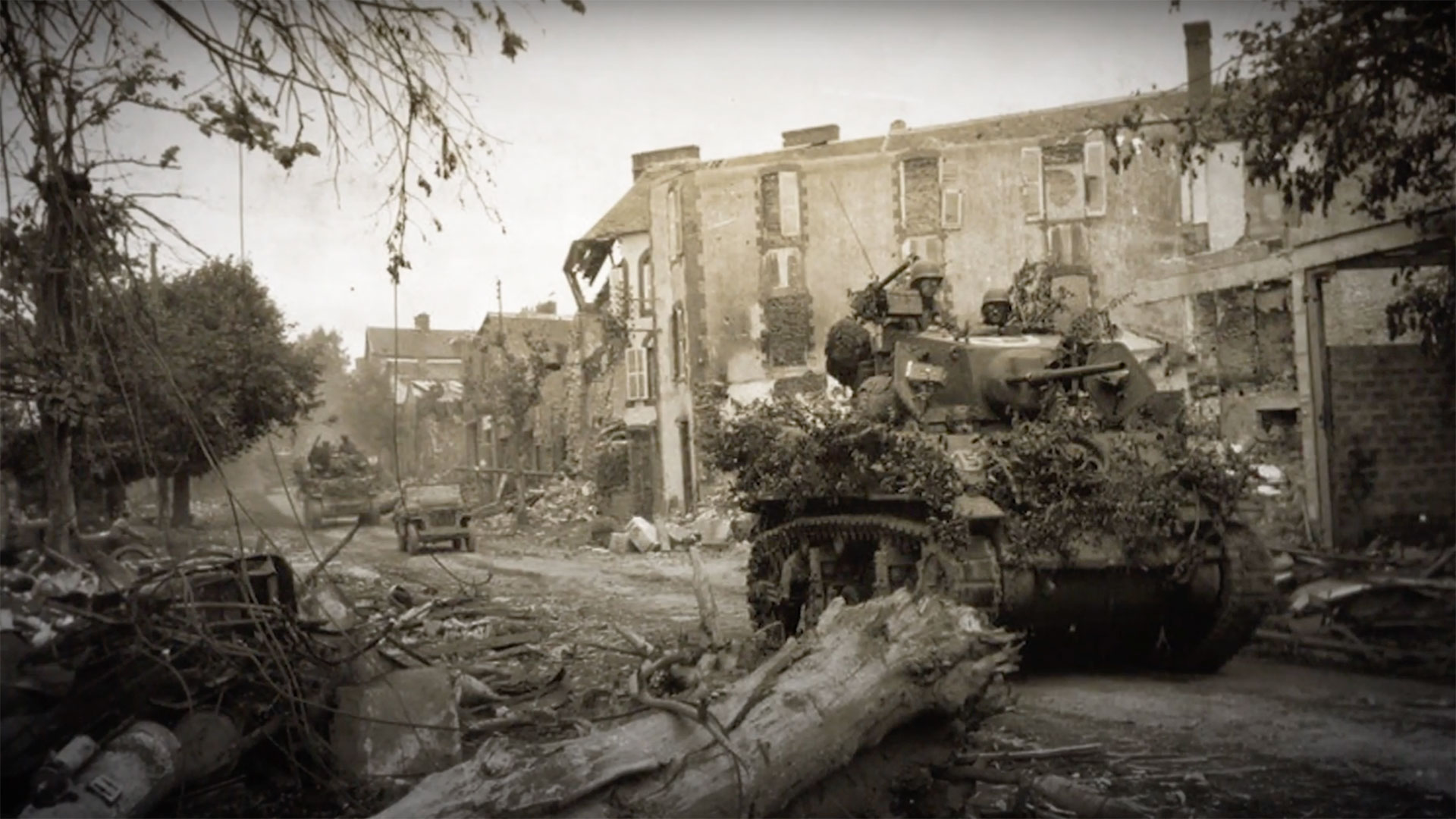 A U.S. M5 Stuart light tank advancing through a French town in the Bocage region. Note the foliage added to the tank by the crew to help make it less visible in the hedgerows.
