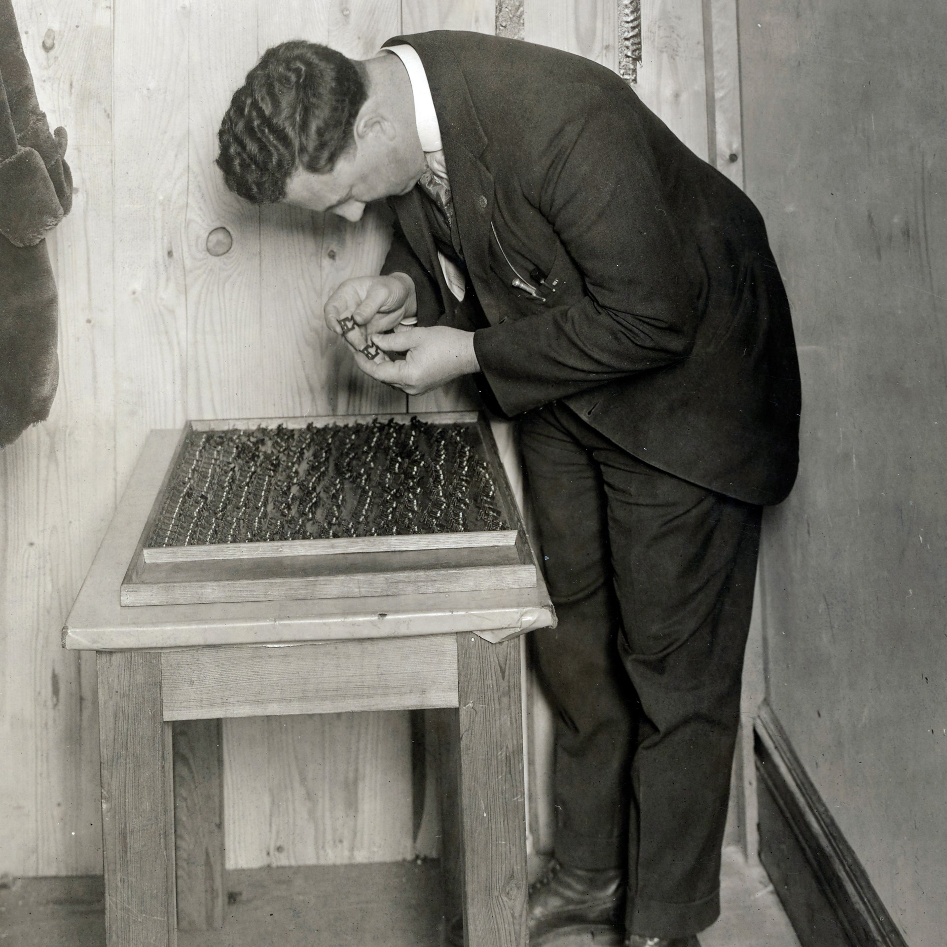 1918 photograph of man in suit hunched over inspecting luminous gun sights on wood table wood walls fur coat hanging