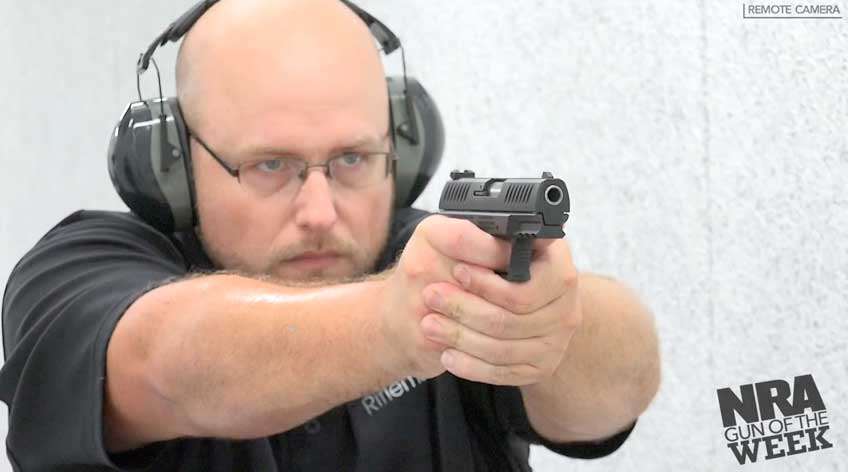 Man in black shirt and wearing protective shooting gear on gun range with pistol in hand.