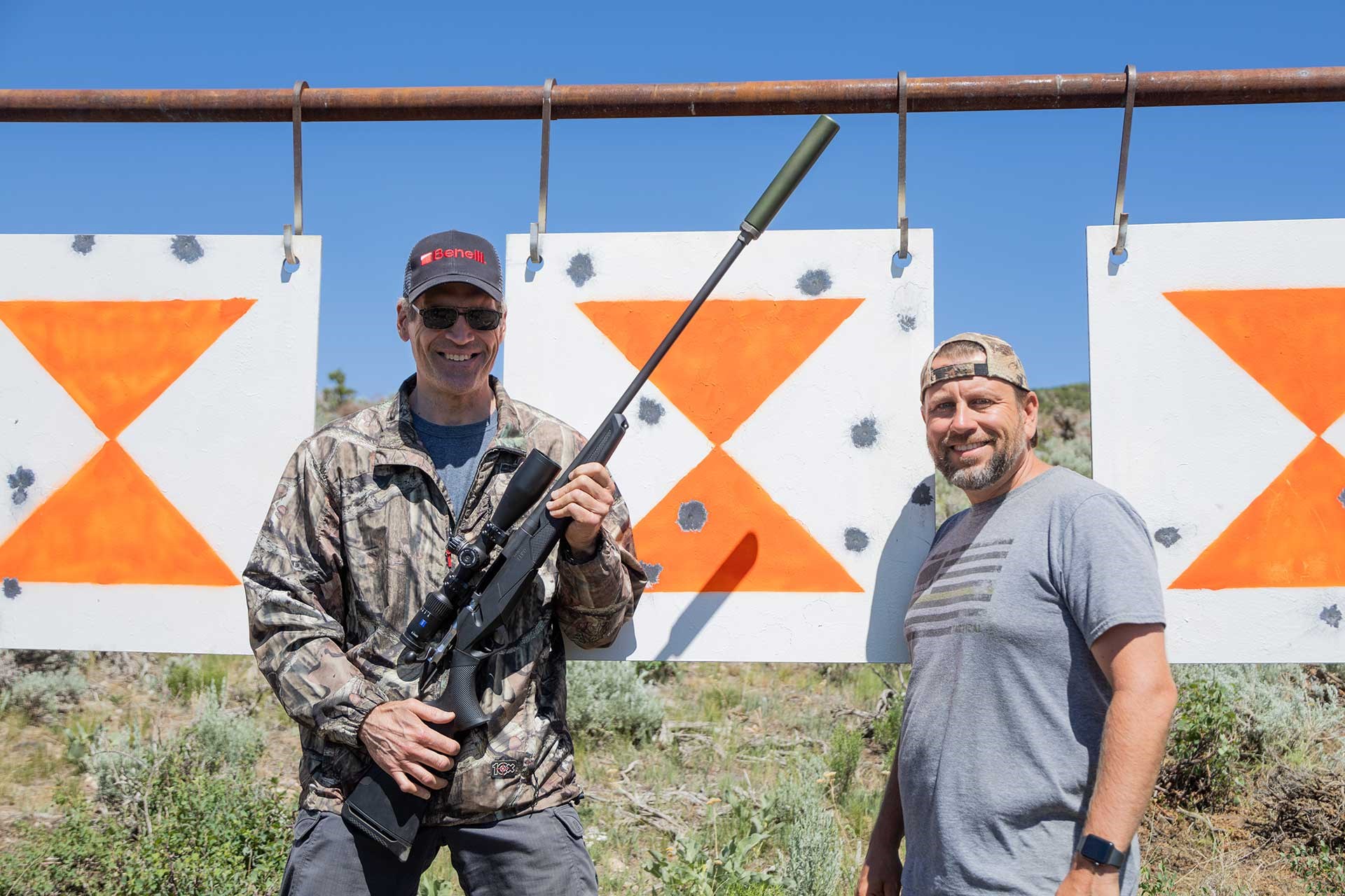 Two men stand next to a steel target showing impact marks. One man is holding a Benelli Lupo rifle with a mounted suppressor.