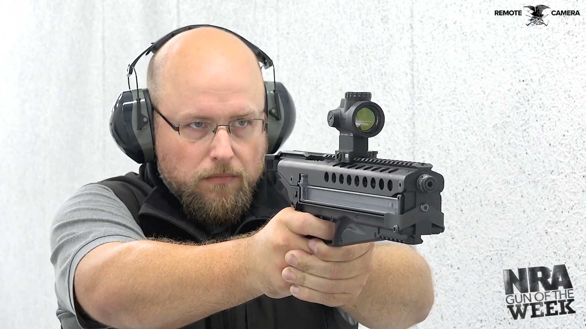 man holding gun earmuffs pistol black metal shooting range white walls text on image noting "remote camera nra gun of the week"