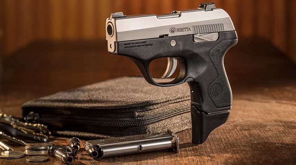 A Beretta Pico standing up on a wooden table, with ammunition, a loaded magazine and a black gun case in the background.