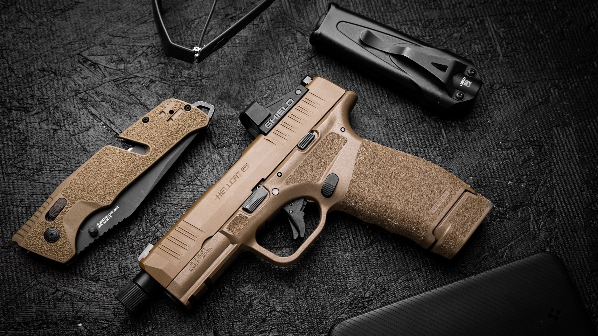 Left side of the Springfield Armory Hellcat Pro OSP in flat dark earth, shown on a dark background next to a knife and flashlight.